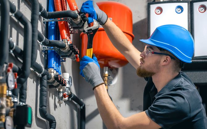 Plumber in a blue hard hat and gloves tightening a pipe with a wrench near water pipes and a red tank.
