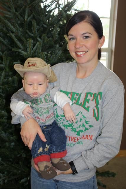 Woman holding baby wearing cowboy hat and Steele's Tree Farm shirt, in front of a Christmas tree.