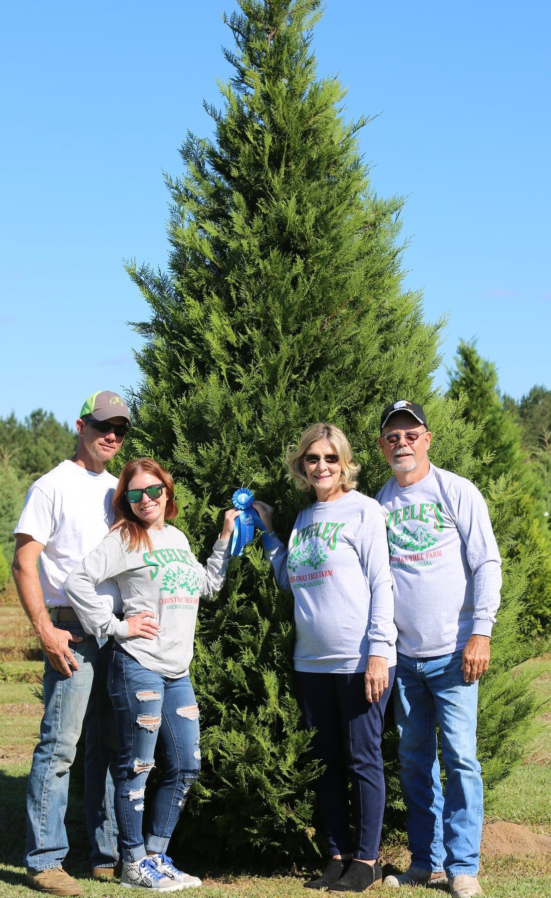 Family poses with a tall, green Christmas tree outdoors under a blue sky; wearing matching long-sleeved shirts.