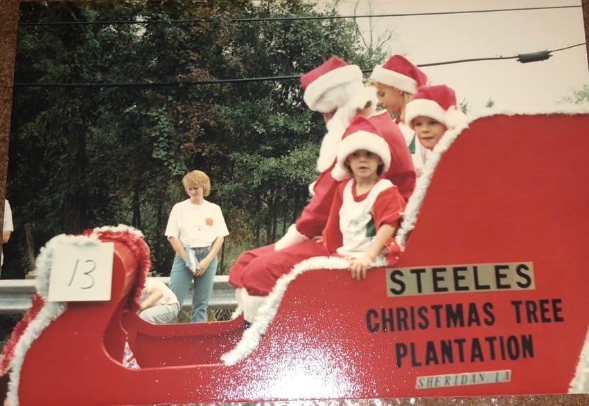 Santa and helpers on a red sleigh float for Steele's Christmas Tree Plantation in a parade, trees in background.