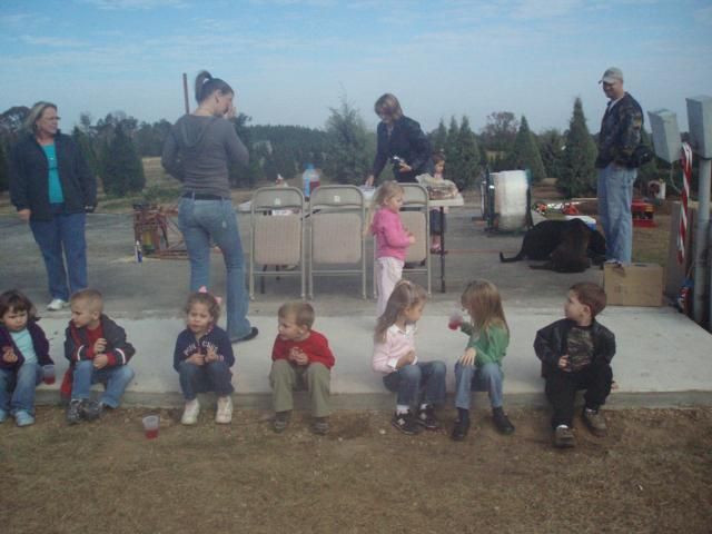 Group of children sitting on a ledge, adults behind a table outside on a sunny day.