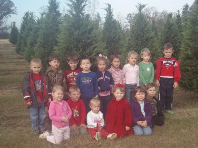 Group of children posing in front of evergreen trees; some are standing, others kneeling.