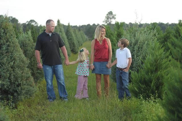 Family of four holding hands, posing in a Christmas tree farm, surrounded by evergreen trees.