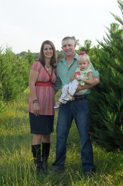 Family posing outdoors with child. Woman in pink top and skirt, man holding child. Green trees background.