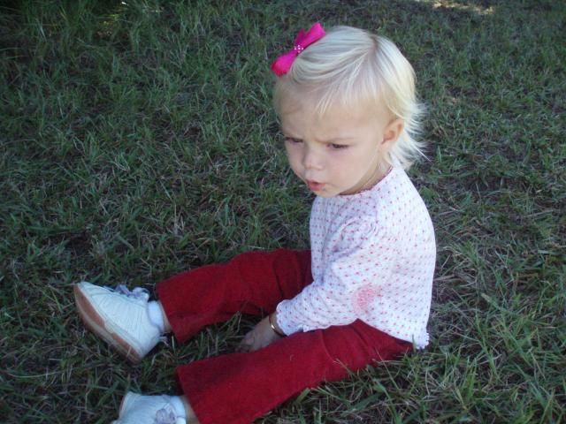 Blond toddler in red pants and white shoes, sitting on grass, with a pink bow.