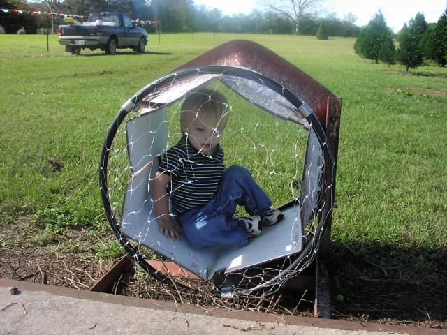 Boy inside a cylindrical play structure with a wire mesh covering, set on grass; a truck is visible in the background.