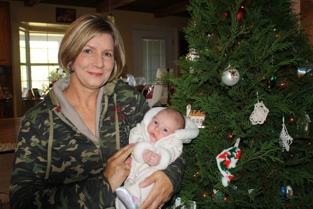 Woman in camo jacket holds a baby near a decorated Christmas tree.