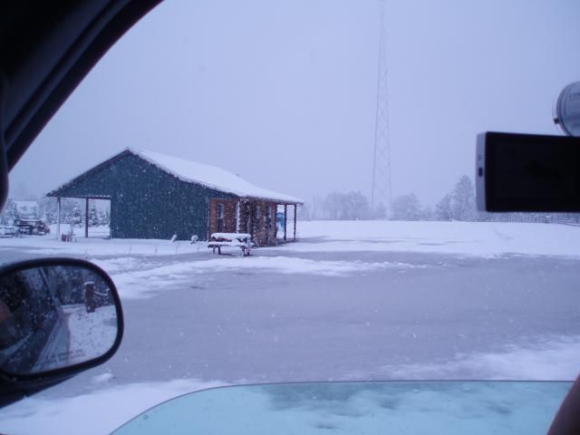 Snowy scene outside a car window: green building, bench, snow-covered field, and a tall tower in the distance.