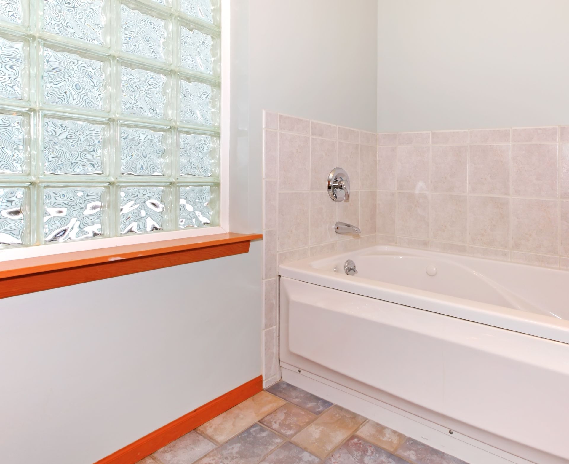 A white bathtub with tiled walls next to a glass block window with an orange-stained wood sill and floor trim. A white bathtub with tiled walls next to a glass block window with an orange-stained wood sill and floor trim.