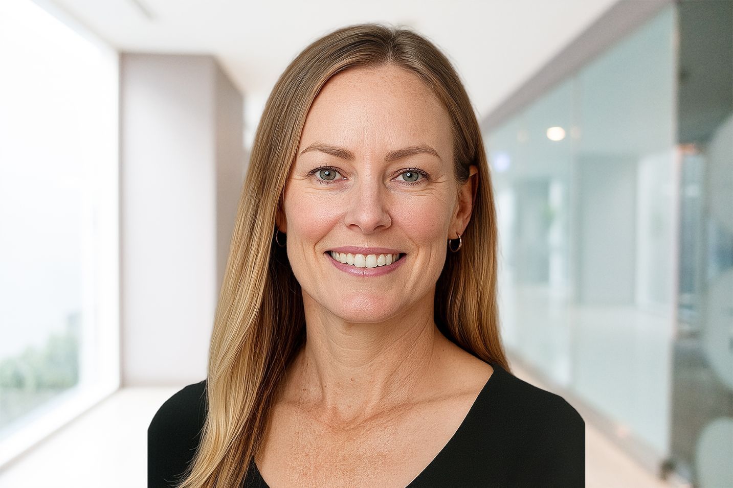 Woman smiling, long blonde hair, wearing a black top. Standing in a bright hallway.