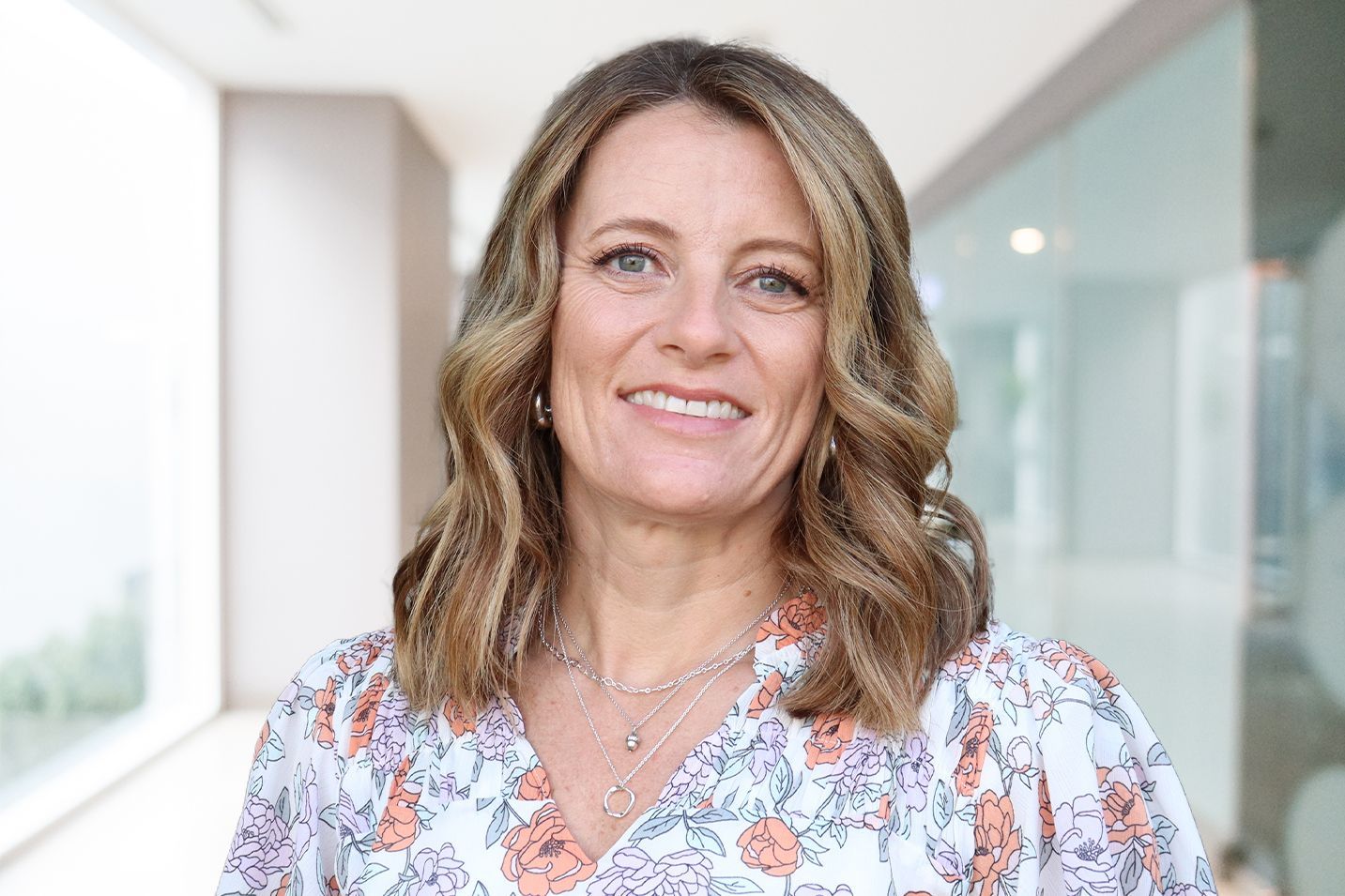 Woman with wavy brown hair smiles in a bright hallway, wearing a floral top and layered necklaces.