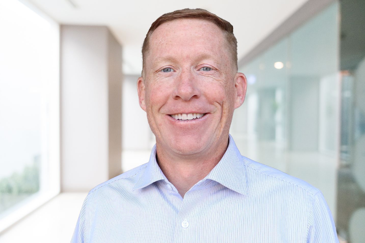Man smiling in a light blue striped shirt, standing in a bright office hallway.
