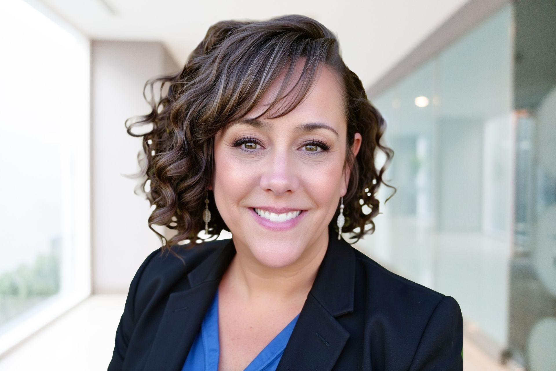 Woman smiling, wearing a black blazer and blue shirt, standing in a brightly lit hallway.