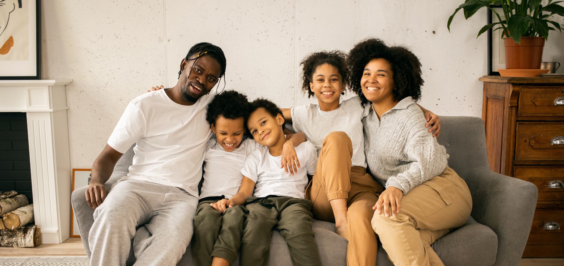A smiling Black family of five sits close together on a couch in a living room.