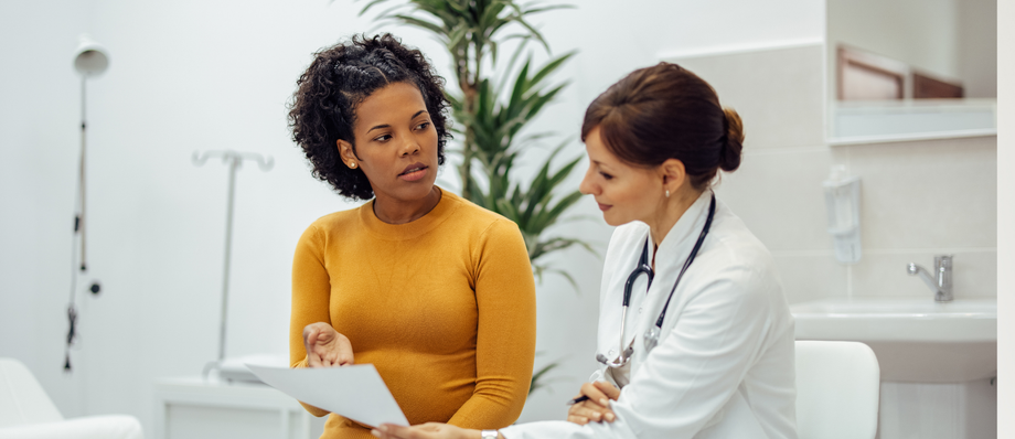 A Black woman in an orange sweater consults with a doctor in a white coat in a medical office.