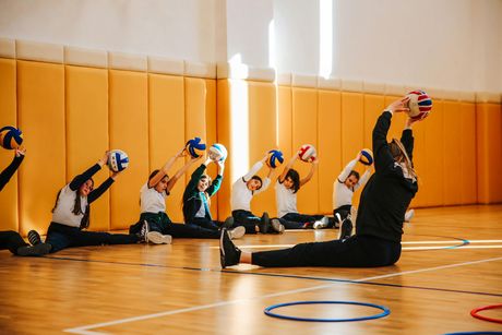 Students and instructor stretching with balls in a gymnasium, extending arms overhead while seated.