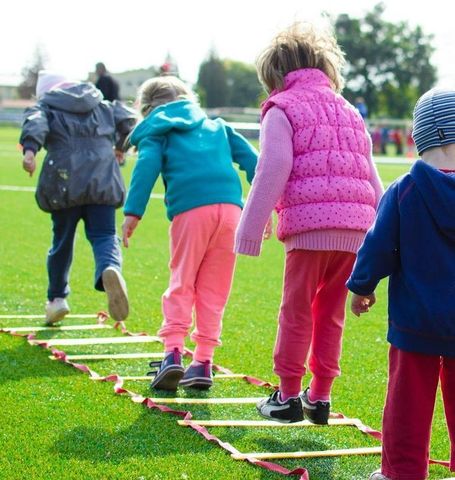 Children running across a red and yellow agility ladder on green grass.