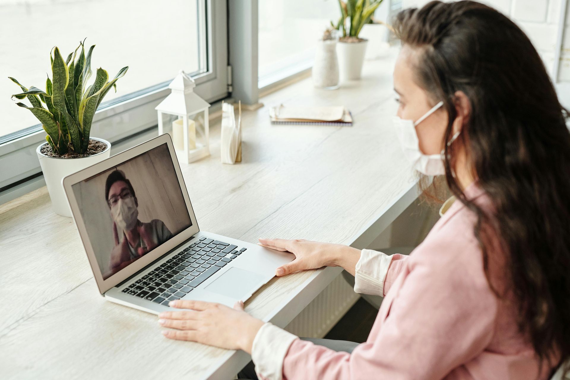 Woman in mask video chats on laptop near window, both wearing masks.