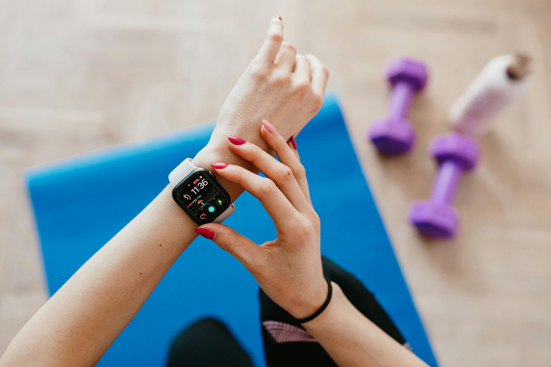 Person checking a smartwatch on wrist, blue mat, purple weights, white bottle.