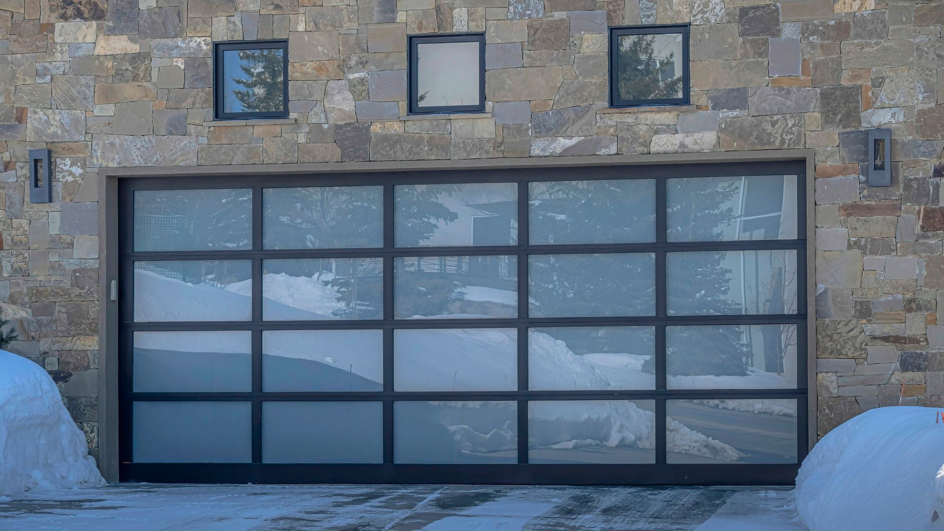 Modern garage door with glass panels, stone wall, three small windows above. Snow present.