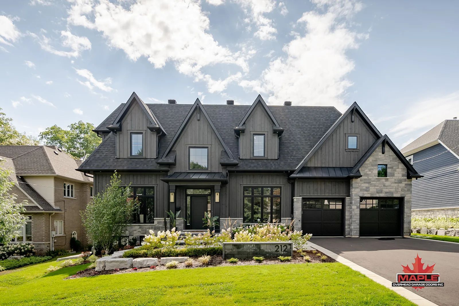 Large dark-gray house with steep gables, black garage doors, and a landscaped front yard under a blue sky