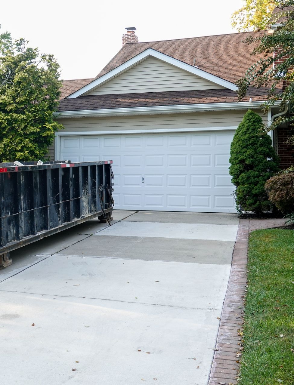 Black dumpster on a concrete driveway in front of a white garage door. House with a brown roof in background.