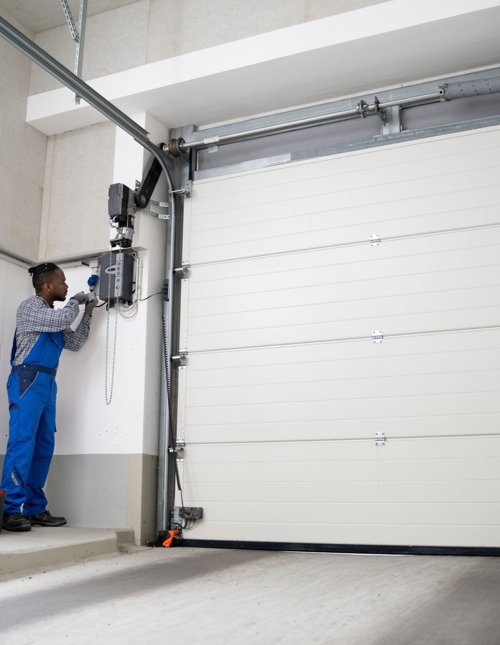 Man in blue overalls repairs a white garage door opener in a garage.