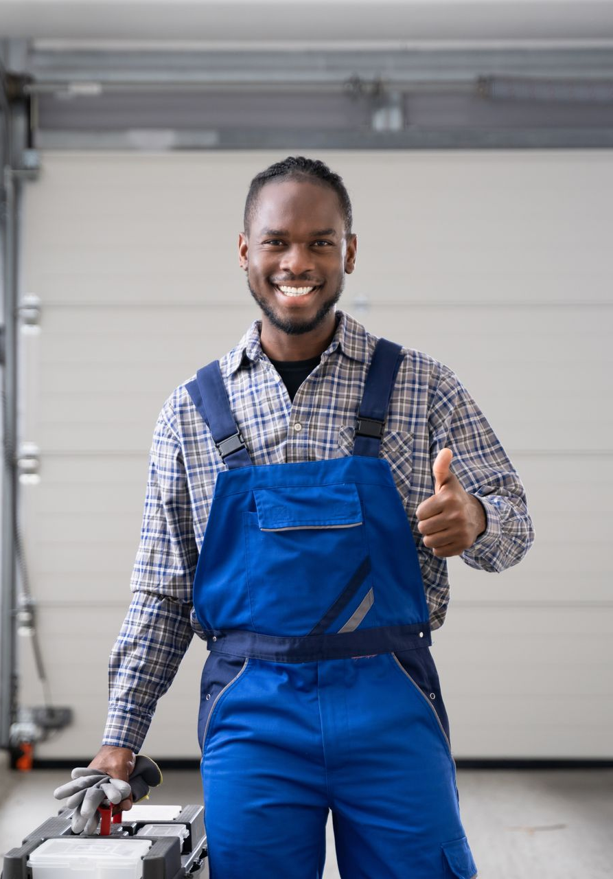 Smiling man in blue overalls, thumbs up, in a garage.