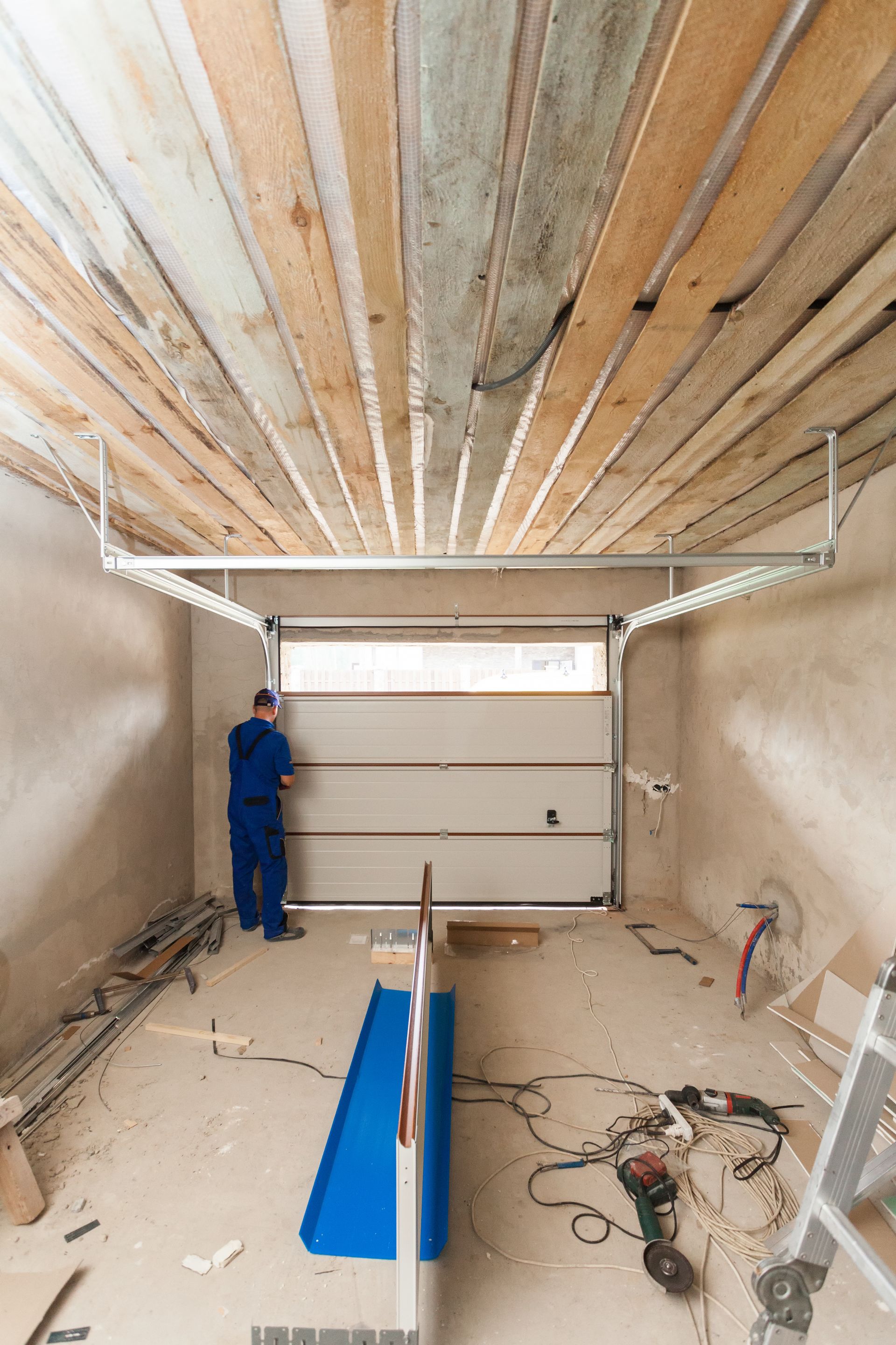 A worker in blue coveralls installing a garage door in a construction setting with exposed wood ceiling.