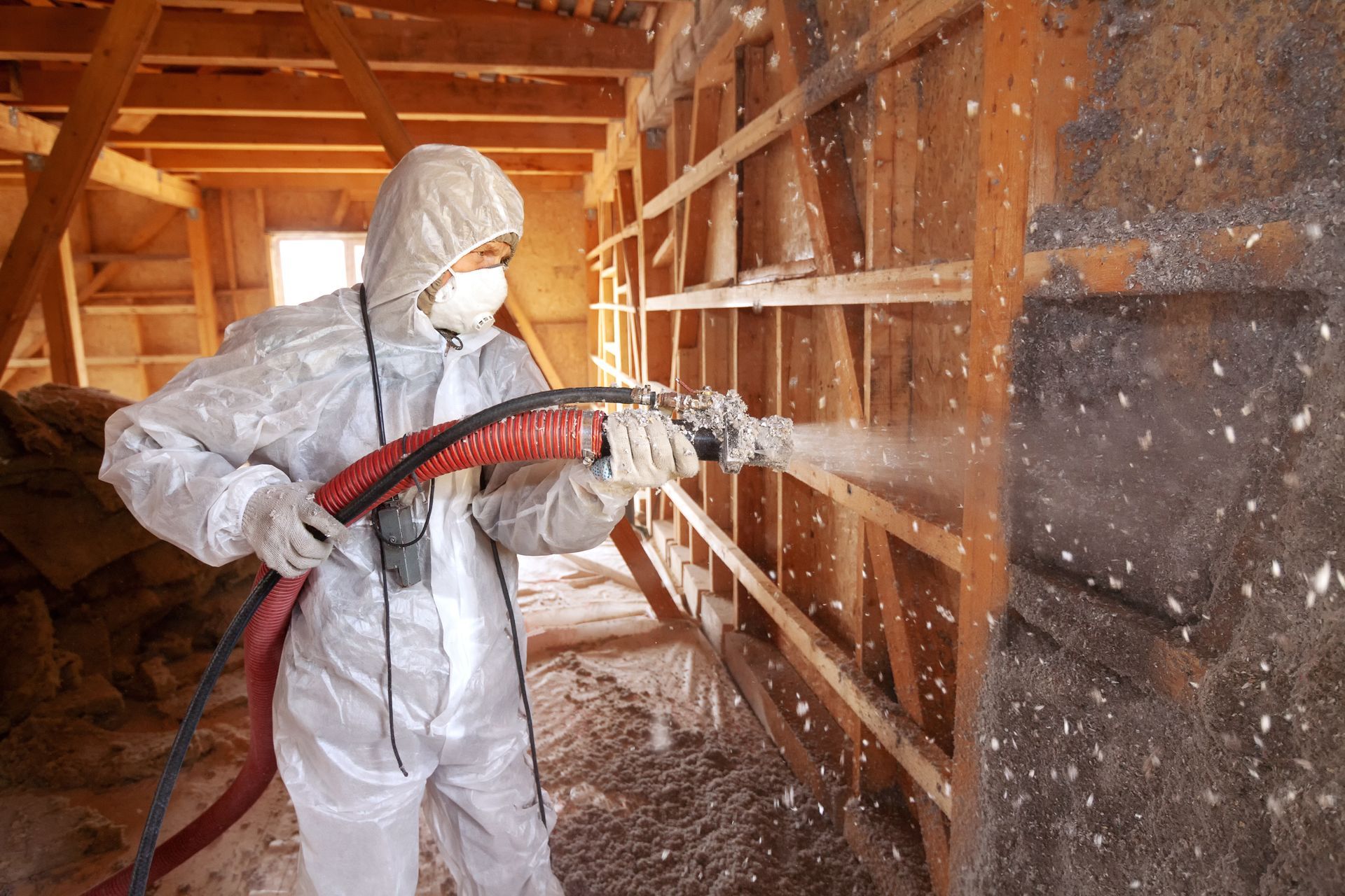 A man in a protective suit is spraying insulation on a wooden wall.