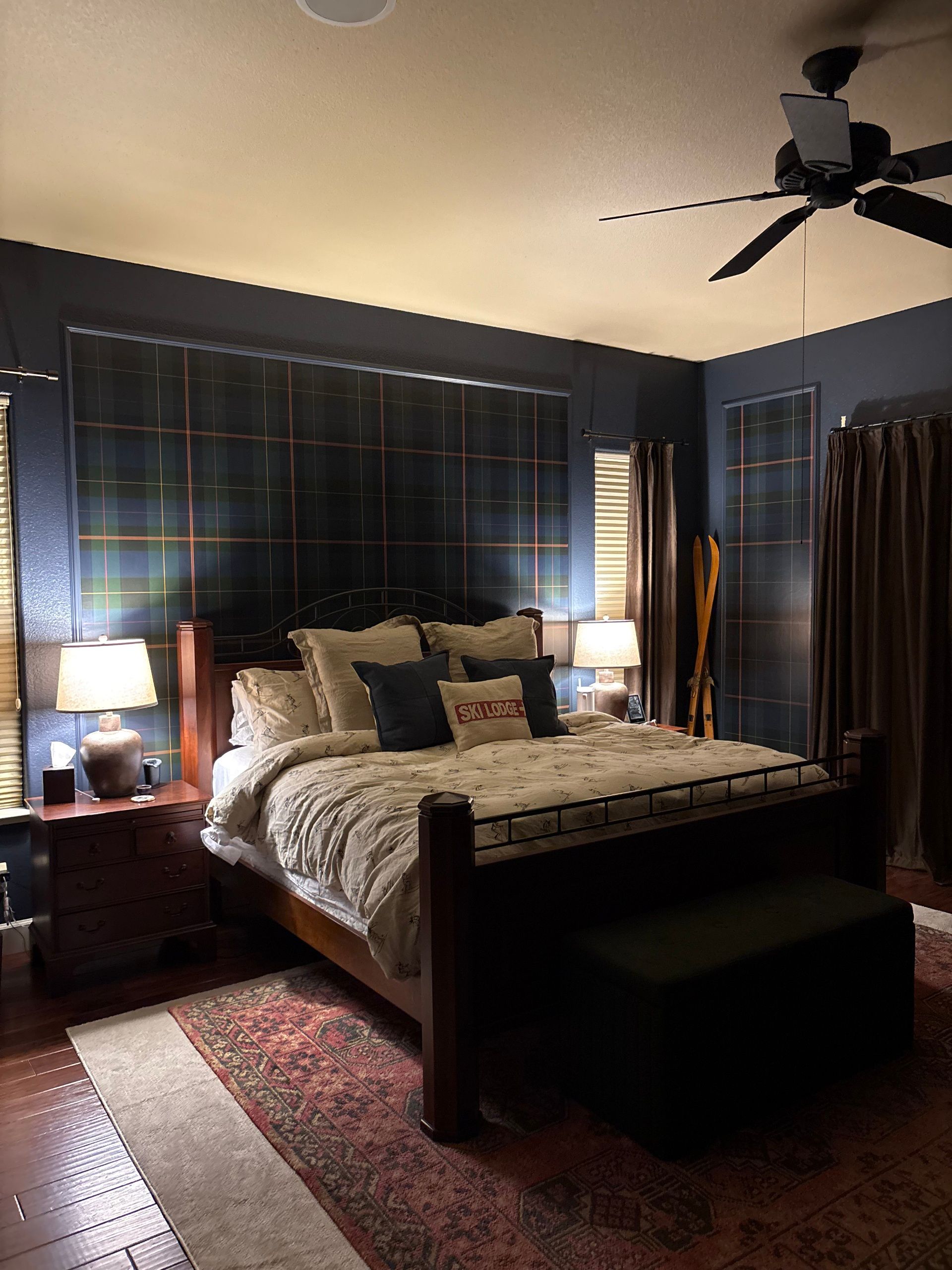Bedroom with a dark blue wall behind a wooden bed, lamps, and rug. A dark-colored bench sits at the foot of the bed.