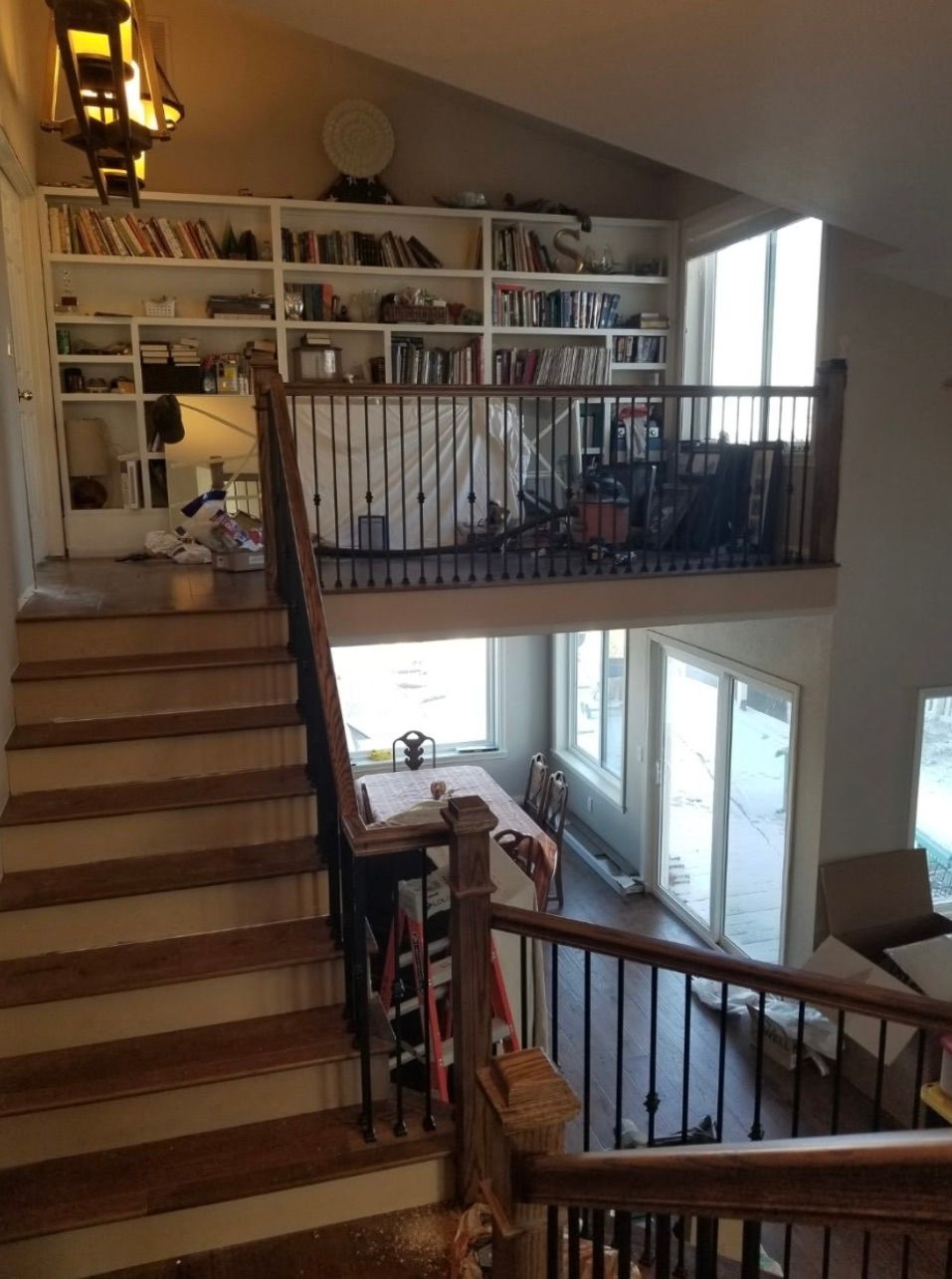 A wooden staircase leads to a loft with bookshelves, and a dining area below with large windows.