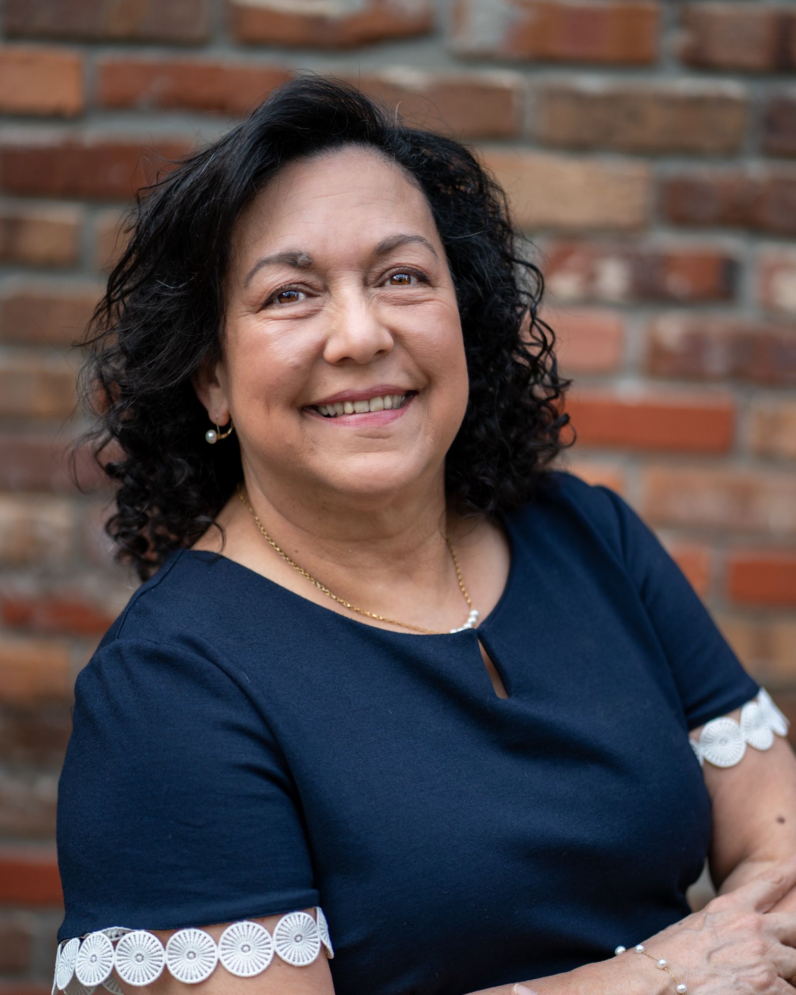 Woman with dark hair smiles, wearing a navy blue top with white scallops , in front of a brick background