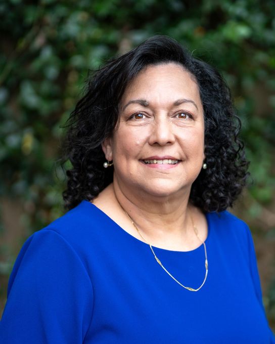 Woman with dark hair smiles, wearing a blue top , in front of a blurred leafy background