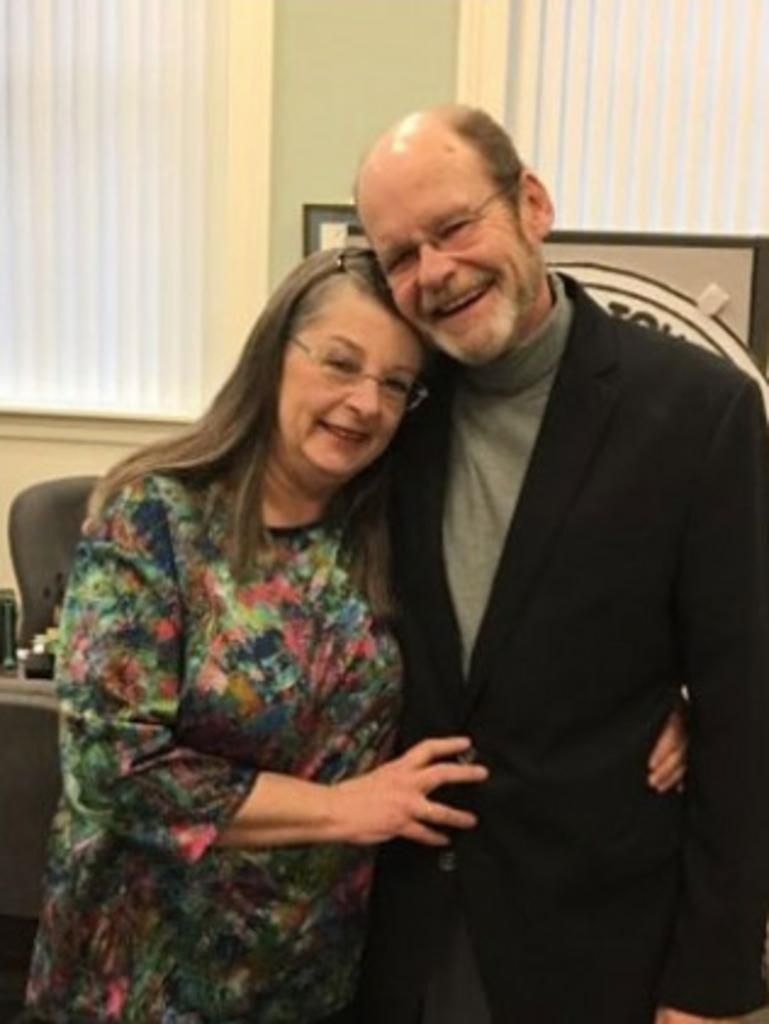 A smiling couple embraces. The woman wears glasses and a patterned shirt, and the man wears a black suit.