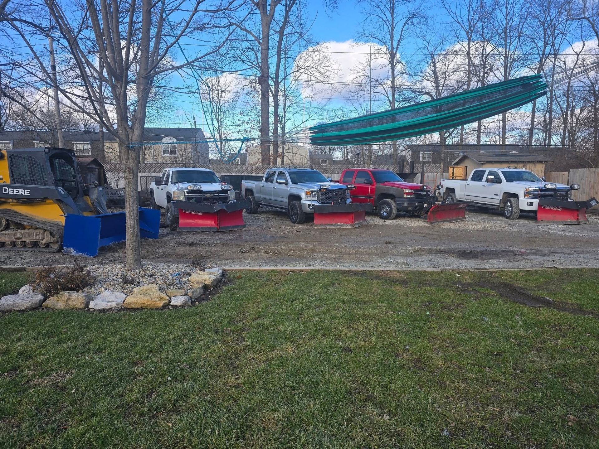 A row of pickup trucks with plows, a skid steer, and a large green structure in a yard.