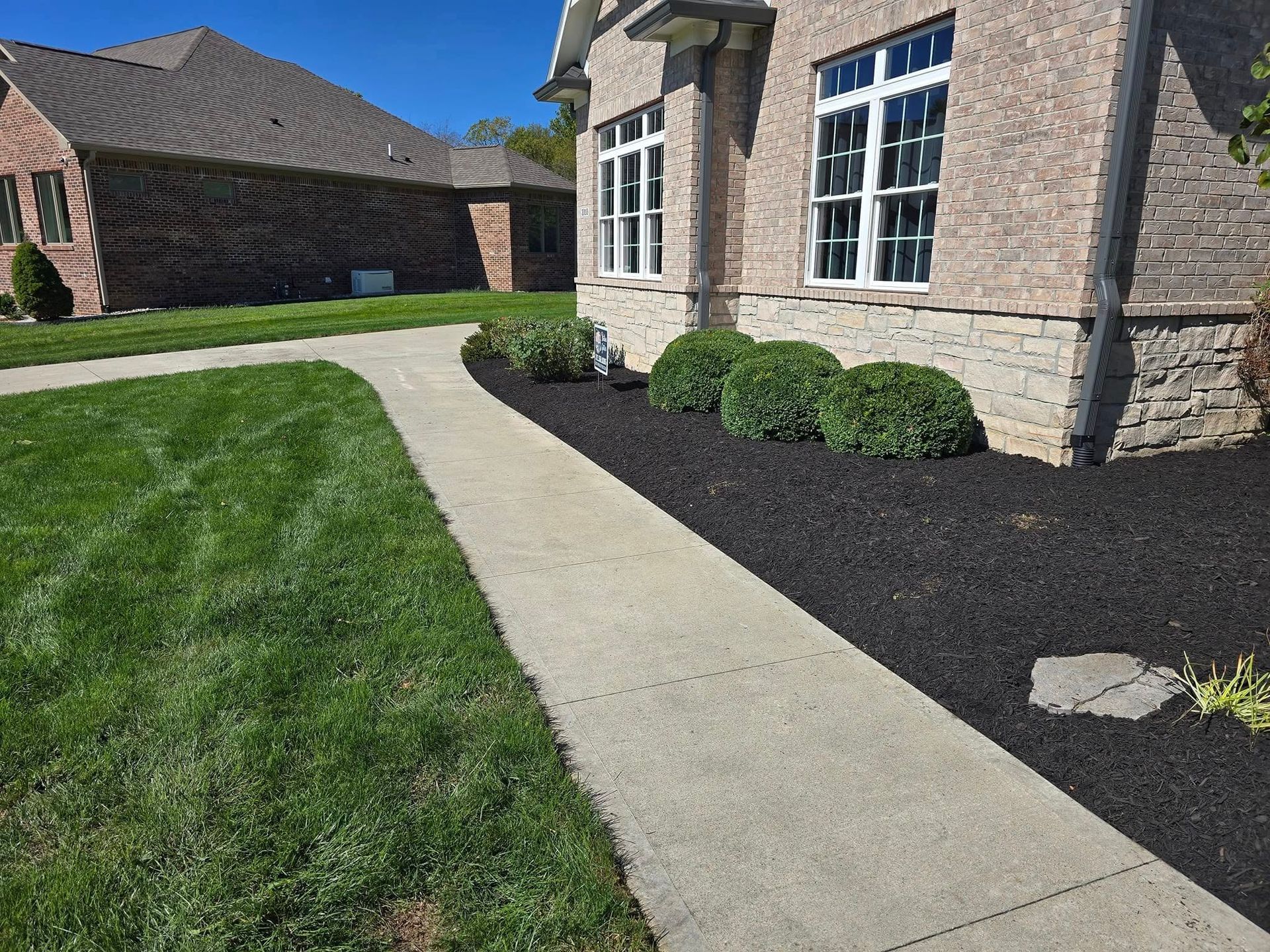 A concrete sidewalk curves along a house with brick and a black mulch bed.