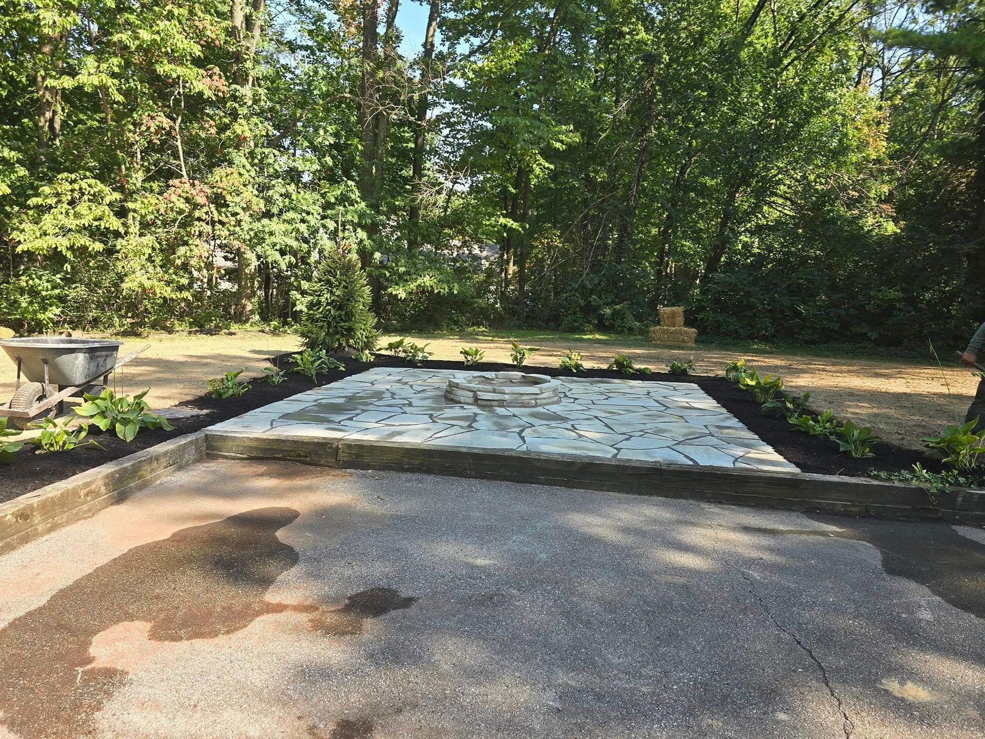 A gravel driveway leads to a stone patio with mulched flowerbeds, surrounded by trees.