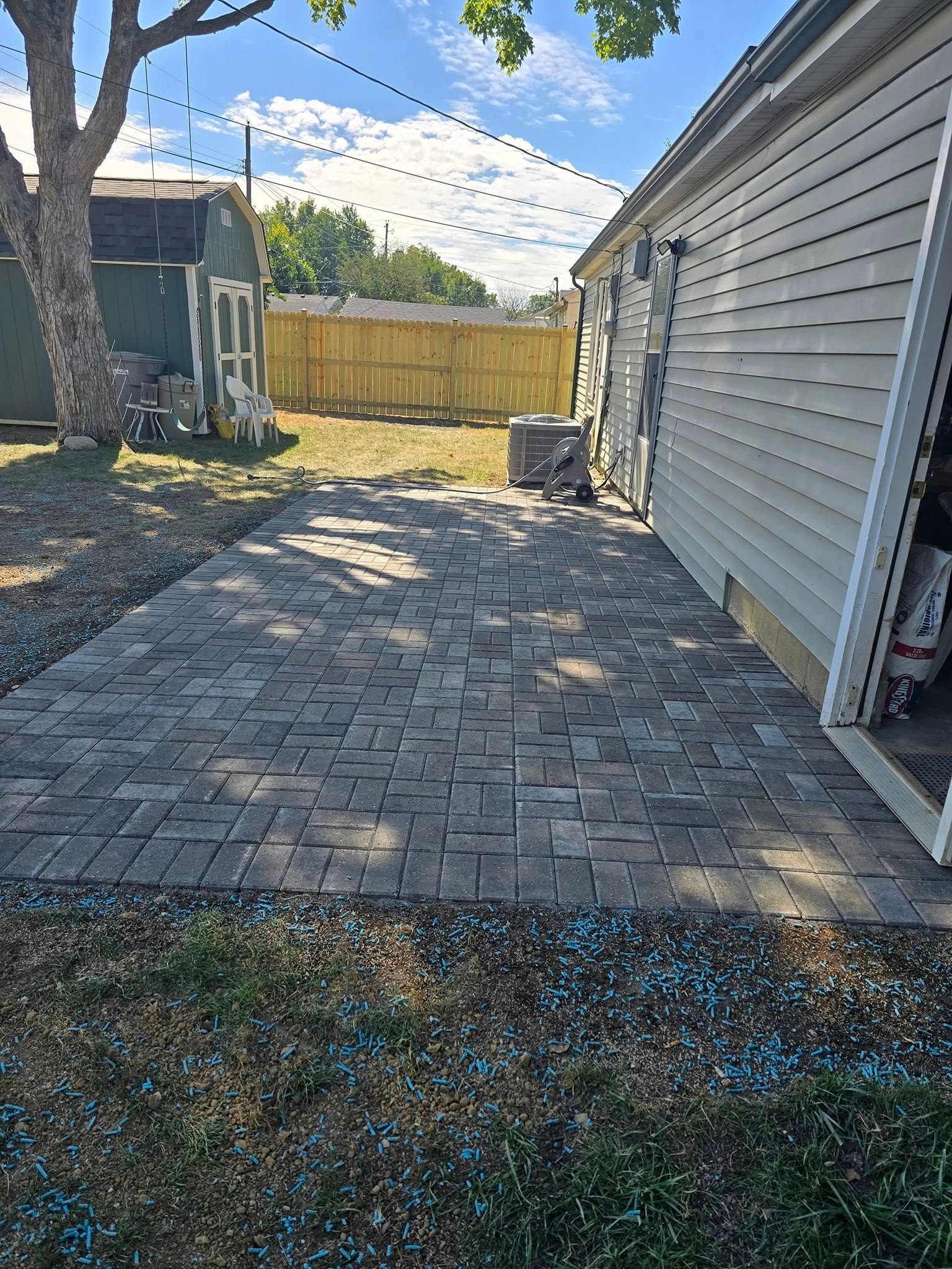 Brick patio next to a house with a green shed and yellow fence in the background. Sunny day.