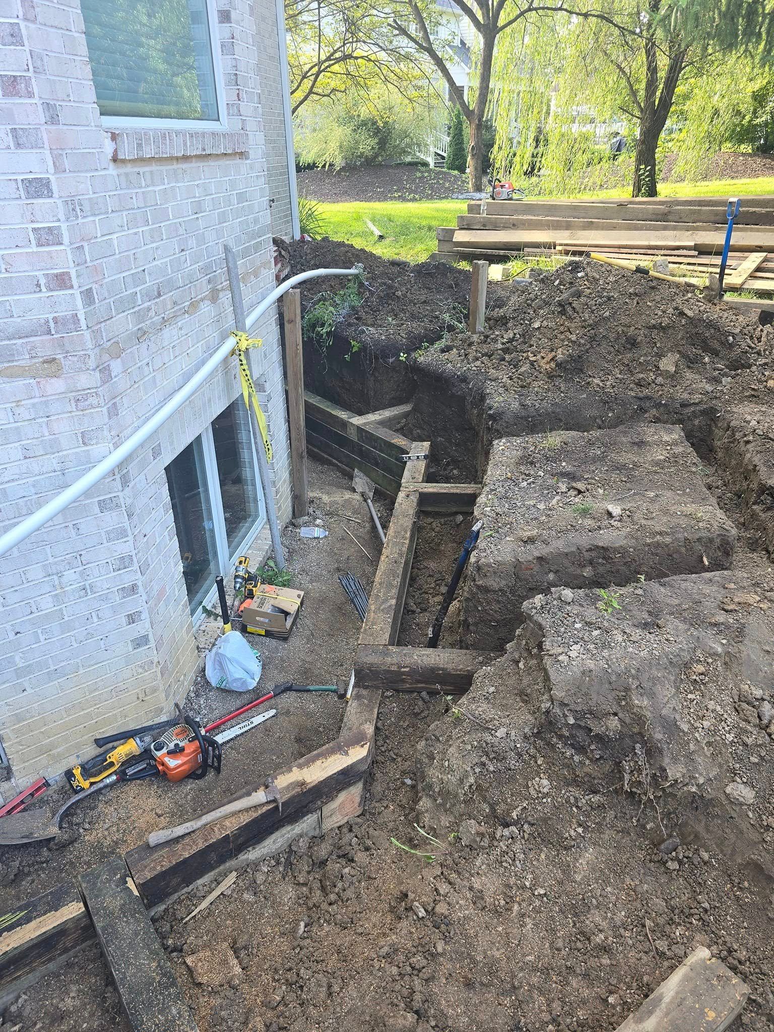 Trench dug alongside a brick building. Construction materials and tools are visible.