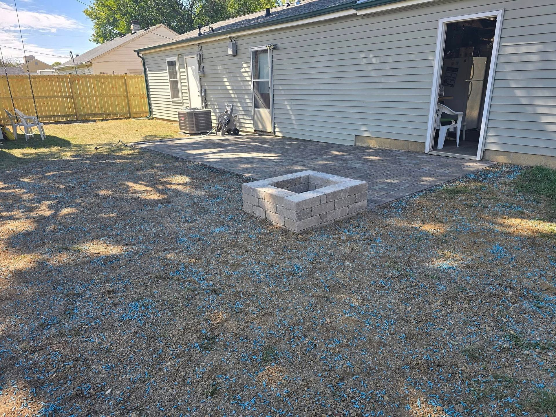 Backyard with gravel ground, a fire pit, and a small patio area. A building with an open doorway is in the background.