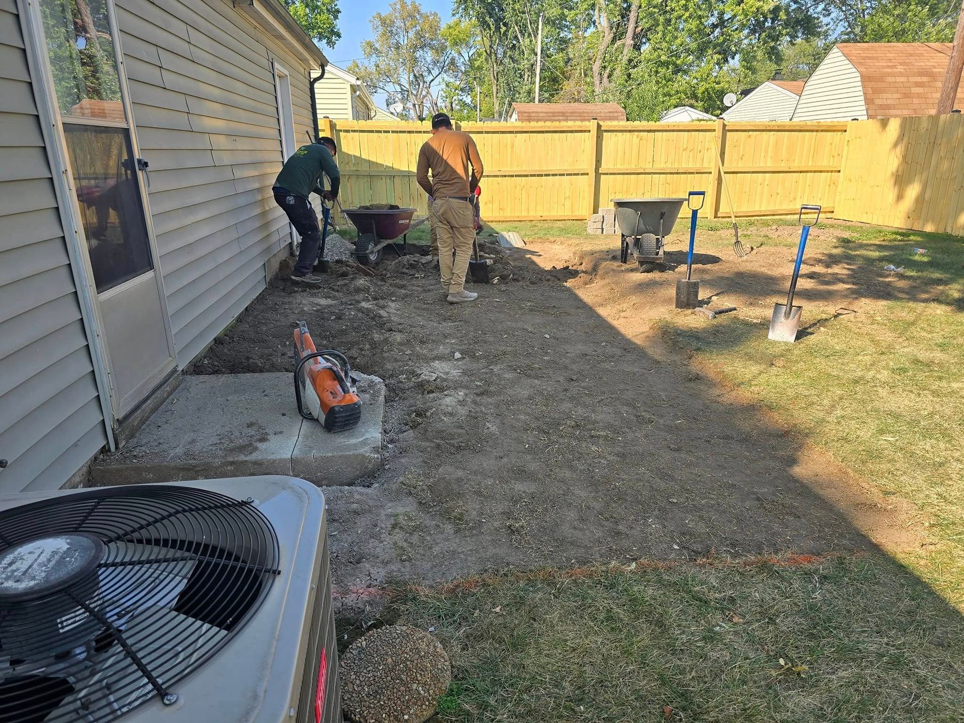 Two workers laying gravel for a patio near a house, with tools and a wheelbarrow in a yard.