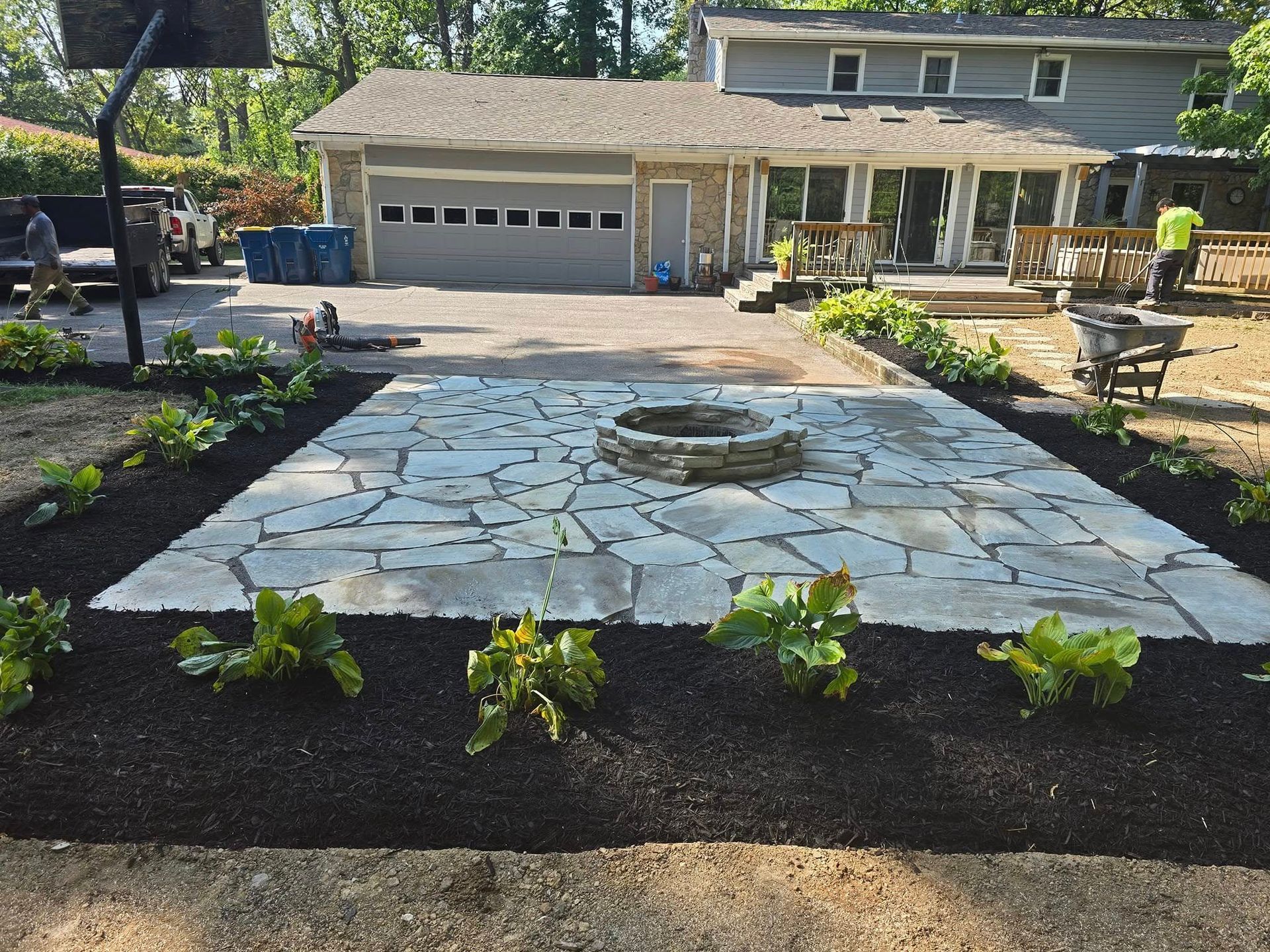 Newly constructed stone patio with a fire pit, surrounded by mulch beds and plantings, leading to a house with a garage.