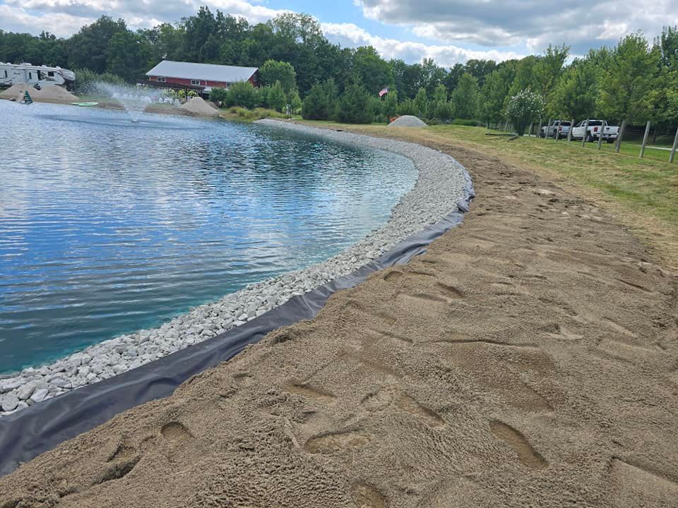Lake shore with gravel edge, black liner, and brown dirt. Trees and buildings in the background.