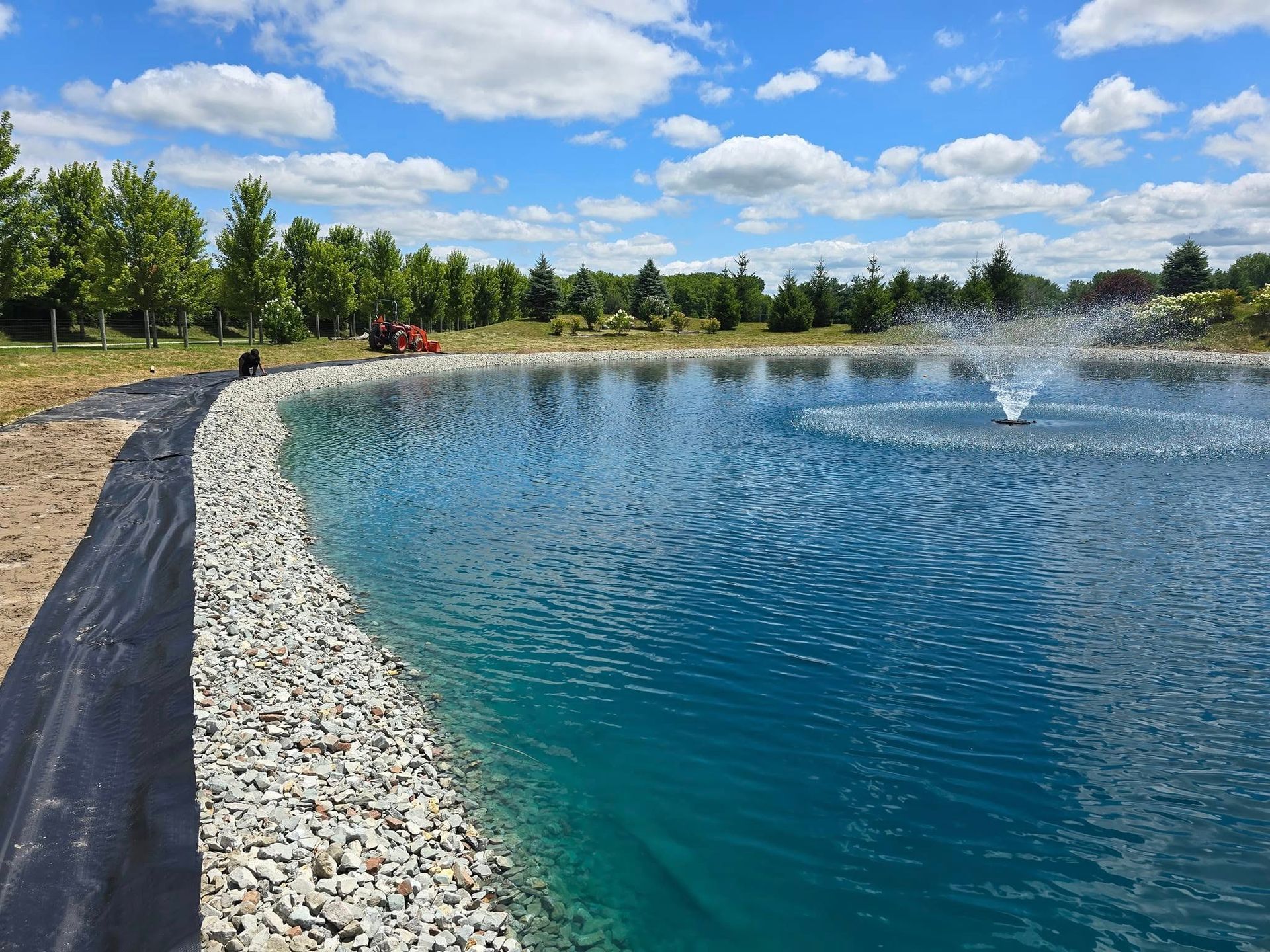 Pond with fountain spraying water, bordered by gray rocks, blue water, and blue sky with clouds.