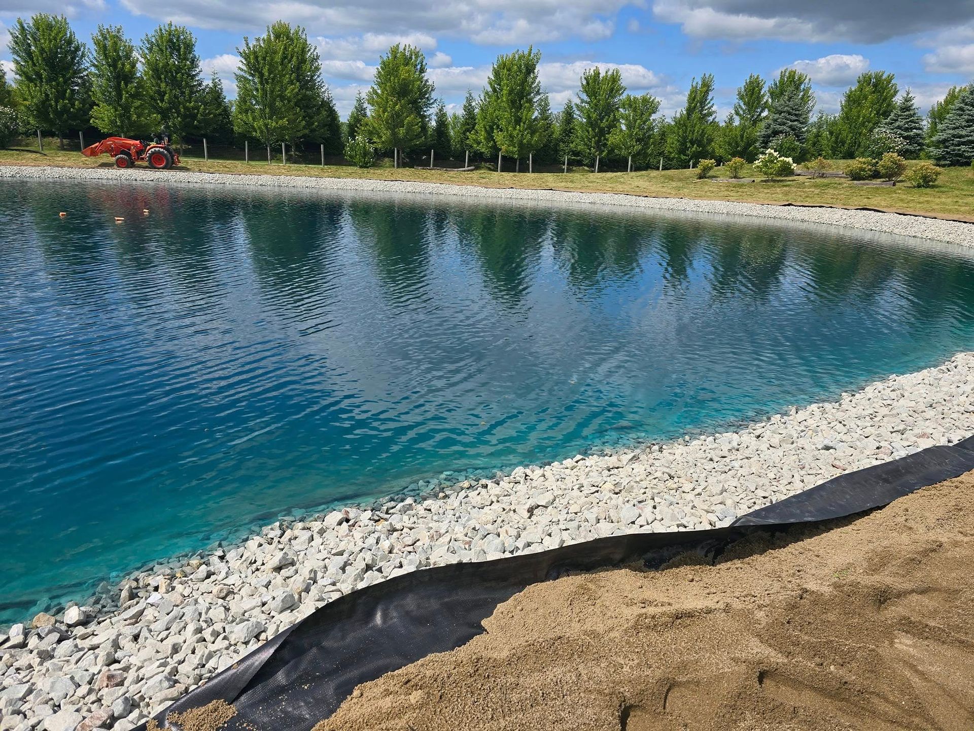 Blue pond with stone edge, green trees, and blue sky.