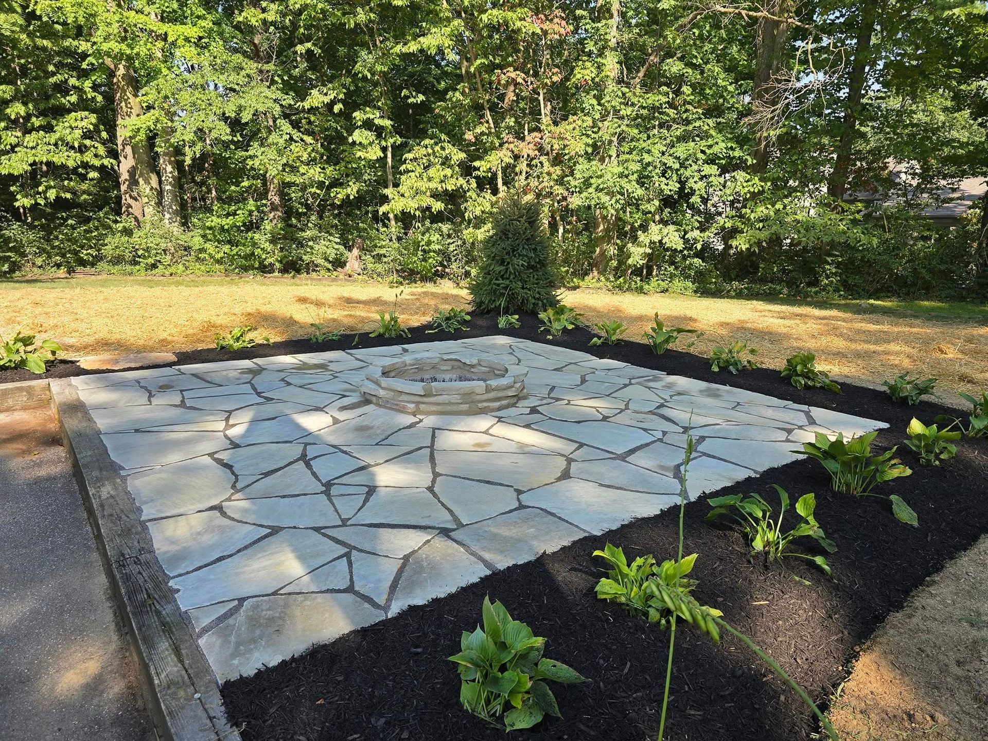 Flagstone patio with fire pit, surrounded by mulch and greenery, against a backdrop of trees.