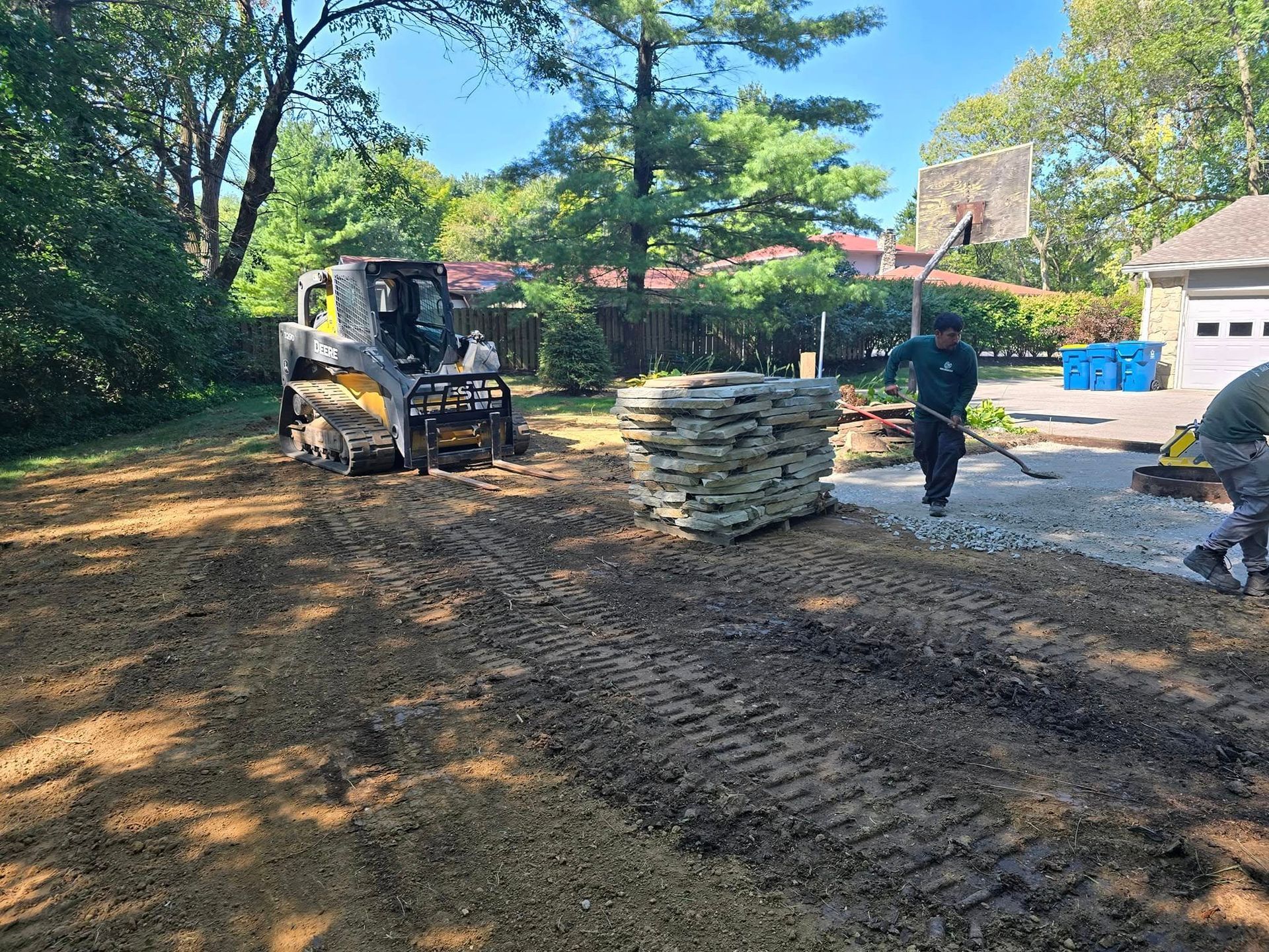 Construction workers laying pavers in a backyard with a skid steer, stones, and trees.