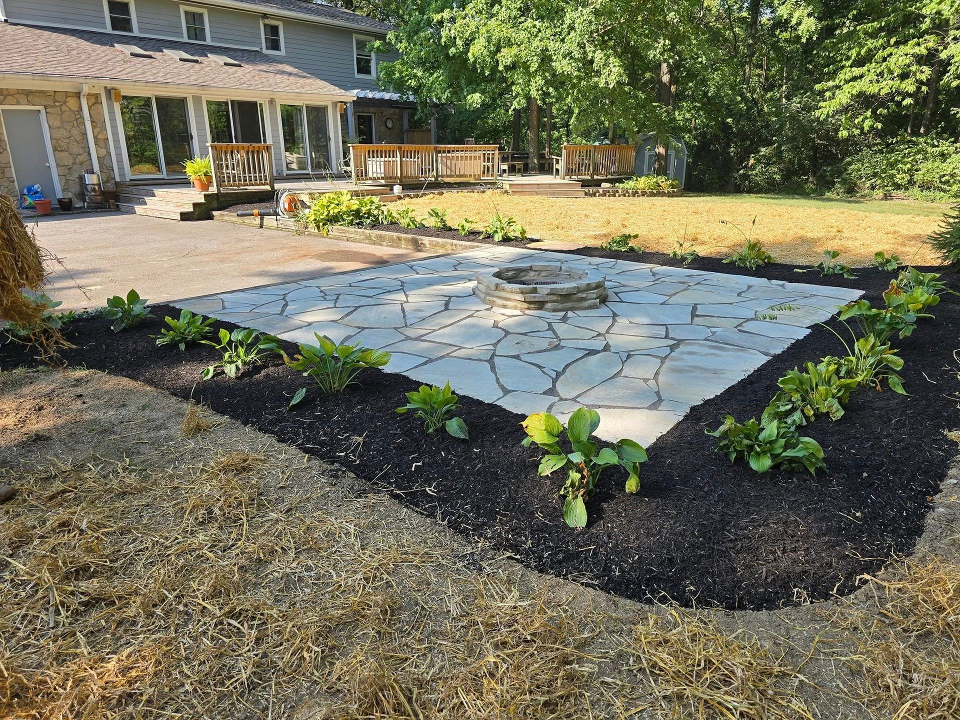 A stone patio with a fire pit surrounded by fresh mulch and young plants in a backyard.