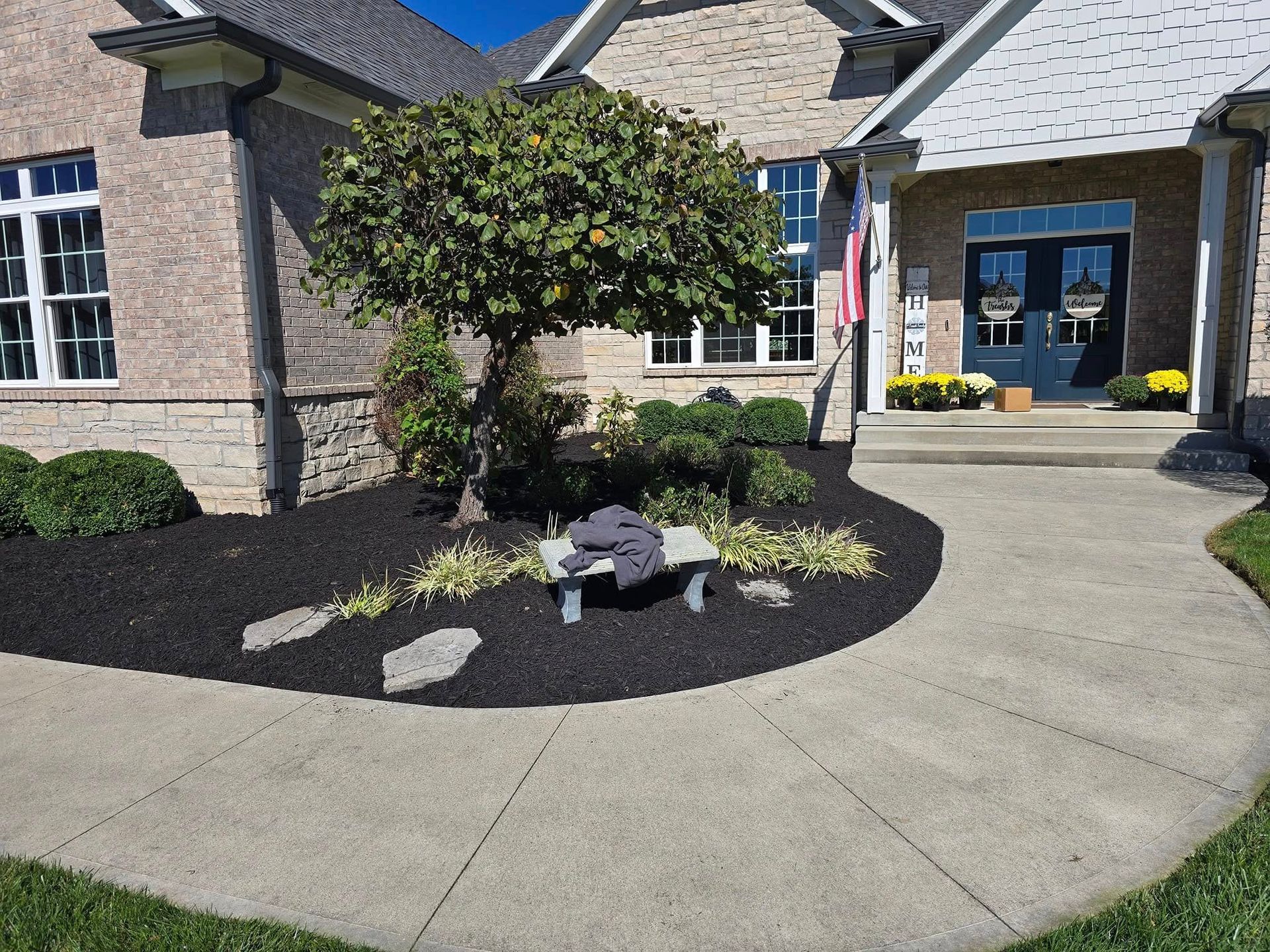A brick house with a curved concrete walkway, black mulch flower bed, a small bench, and a tree.